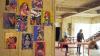 Colourful artwork depicting powerful women which is  displayed on a wooden wall. In the distance are two female artists working at a table