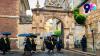 Students in graduation robes walk through a stone archway holding umbrellas.