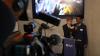 Two young male students stand in front of a video screen holding microphones. There is a British Council branded lectern in front of them. A camera man can be seen filming them in the foreground.