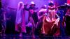 Premiere of ‘Nobody Whistles Anymore’ in Ljubljana, May 2023. Visual description: On the darkened stage a crowd of people in colourful costumes is dancing. At the front of the stage: a person in pink dress with a pink crown, a male in drag and a women with metal helmet, military jacket and fishnet stockings. Flower petals on the floor and some smoke in the background.