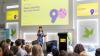 Emily Kasriel on stage talking to an audience. A British Council branded video screen can be seen in the background saying: Deep Listening with Emily Kasriel.