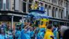 Photo from The Blue and Yellow Submarine Parade in Liverpool. The photo shows a big submarine-like parade float with glass windows. It is painted in colours of the Ukrainian flag (blue and yellow), and there are performers inside and on top of the submarine, performing. In the foreground, parade participants, visitors and revellers are dancing and celebrating in blue and yellow outfits. 