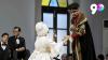 An actor and an actress on stage in period costumes. The actress is wearing a white dress and a white wig and the actor is wearing a black hat and a fur trimmed robe. Two actors in dinner jackets are watching from one side.