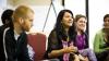 A woman in the centre of the image is talking to her peers in a group discussion during a worksho. Two men are listening to her on her left and two women are listening to her right. There is a flipchart in the background.