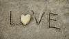 The word 'love' written in wet sand, with a heart shaped stone for the letter 'o'.