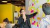 A young woman inside pointing to a whiteboard with colourful stick-on notes, explaining work processes to another male colleague in the office