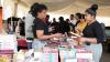 In the background: a room busy with people looking at and buying books. Close up: a bookseller talks to a woman holding one of the books from the display. 