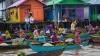 Indonesian women rowing in boats down a river carrying foodstuffs against a background of colourful houses on stilts on water.