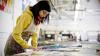 A young woman wearing a white apron covered in paint splatters looking at a selection of art works laid out on a large table in an artist’s studio