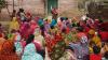 Woman addressing a large group of women and children gathered outdoors in a rural community setting.