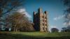 The large tower of Penrhyn Castle in North Wales, constructed in the style of a Norman castle. It has large turrets on each corner and is set in grass surrounded by wintry trees.