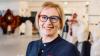 Lady with blonde hair, dark blue top and colourful glasses, smiling.