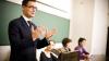 Main in a suit speaking to an audience out of shot. He's standing in front of a podium. Three female panelists are sitting at a table next to the podium, out of focus.