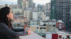 Young woman holding a coffee is looking out at an urban landscape from a rooftop terrace.