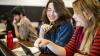Two female students in a lecture theatre are in the foreground, smiling and consulting their laptops. A male and a female student are in the background reading.