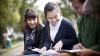 Three students are sitting on a bench outside, smiling and consulting papers that the middle student has on her lap.