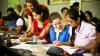 In a classroom full of students, four female students are in the foreground, smiling and studying, writing in their jotters.
