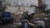 Young boy wearing ripped jeans, padded coat, with the hood of a striped hoodie pulled up, sits in the rubble of a bombed house looking at a book.