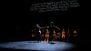 Performance on stage with two performers holding hands and pulling away from each other.