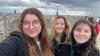 Three young women smile at the camera. Paris and the Eiffel Tower can be seen in the background.