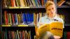Woman sits in a library with shelves of text books behind her. She is holding a book call 'How English works'.