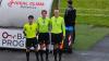 Three football referees in lime green jerseys standing together at the side of a football pitch.