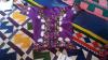 Short sleeved purple jacket with rich decoration around the collar, hem, arm hems and each side of the centre front opening, it is lying on quilted material with geometric patterns.