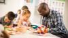 A teacher sitting at a desk helping a child with her work. Another child can be seen at the desk and two more children are in the background.