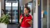 A woman with headphones around her neck and holding a red notebook is walking down a corridor.