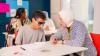 A teacher wearing a hijab is sitting beside a visually impaired student who is wearing sunglasses. They are engaged in a one-on-one learning session at a classroom desk with papers and books in front of them. Other students and colourful posters are visible in the background.