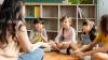 Four primary school students sit on the classroom floor listening to a female teacher.