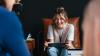 A young woman sitting down on a couch, studying on a tablet. She seems to be chatting with her friend who has a laptop open before her. 