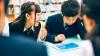 Two Japanese students in school uniform play a game on a tablet.