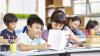 Six young learners are sitting at desks in a classroom. In the front row two young learners are comparing papers.