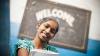 A school girl in India smiling and standing below a 'welcome' sign