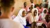 Students in a classroom wearing pink uniforms, with two girls in white headscarves looking toward the front where a teacher is speaking.