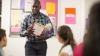 Male teacher standing in a classroom holding a book and talking to pupils
