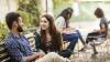 A man and a woman are sat on a bench chatting. A few students are sat on chairs in the background out of focus.