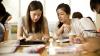 Two female students studying at a table looking over a text book with other students around them.