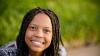 A black female smiling at the camera seated with out of focus grass behind her.