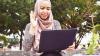 Woman sitting outside with a laptop on her knee. She is waving at the screen and smiling.
