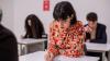 Three adults are sitting at desk in an exam room, taking a writing test