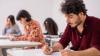 Four adults are sitting at desks in an exam room, taking a writing test