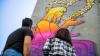 Two people admiring the street artwork of a young female in a wheelchair in bright colours- commissioned as part of the Accessibility Film Festival in Turkey 