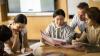 Four young people are sitting around a table, reading, with a teacher sitting behind them